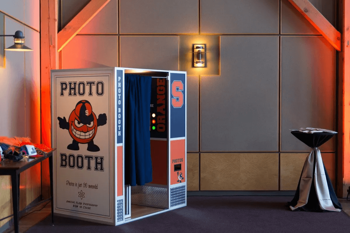 Syracuse Orange wrapped photo booth with Otto panels and a prop table staged beside it in an uplit venue