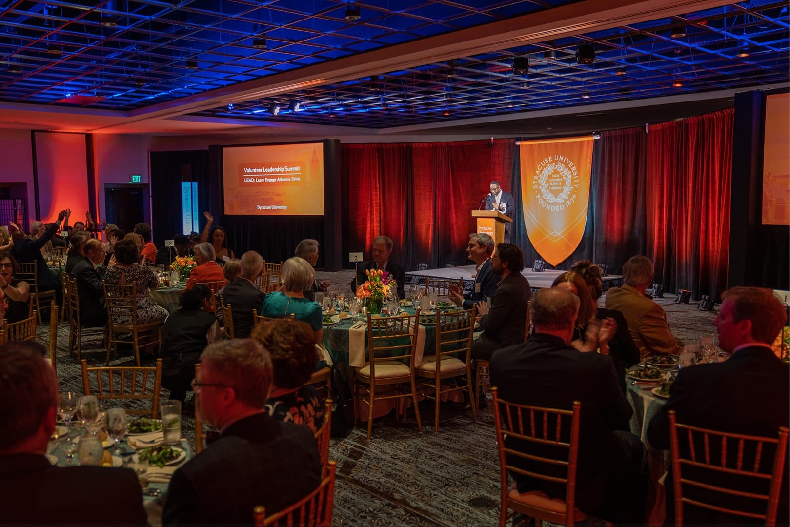 A professionally lit fundraising dinner with a speaker, stage, and projection screens